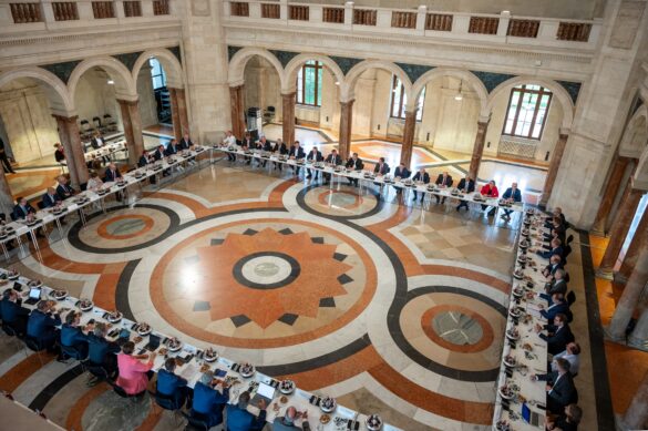 Blick von oben auf eine große Runde von Konferenzteilnehmern an langen Tischen in einem historischen Saal.