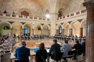 Zahlreiche Teilnehmer sitzen in einem großen historischen Saal an langen Tischen während einer Konferenz.