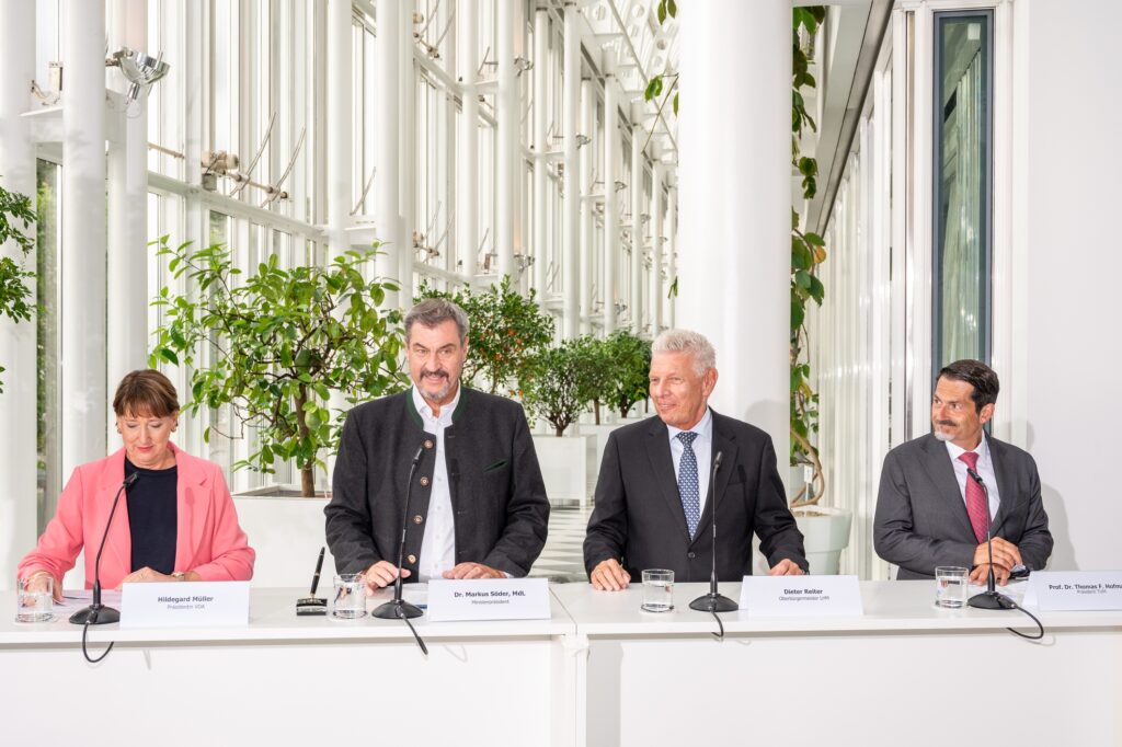 Vier Personen sitzen bei einer Pressekonferenz an einem Podium mit Mikrofonen.
