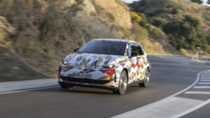 Getarnter elektrischer Kleinwagen fährt auf kurviger Landstraße durch hügelige Landschaft.