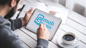 Person nutzt ein Tablet mit dem Logo von E-Mob-Train auf dem Bildschirm, daneben steht eine Tasse Kaffee auf einem Holztisch.